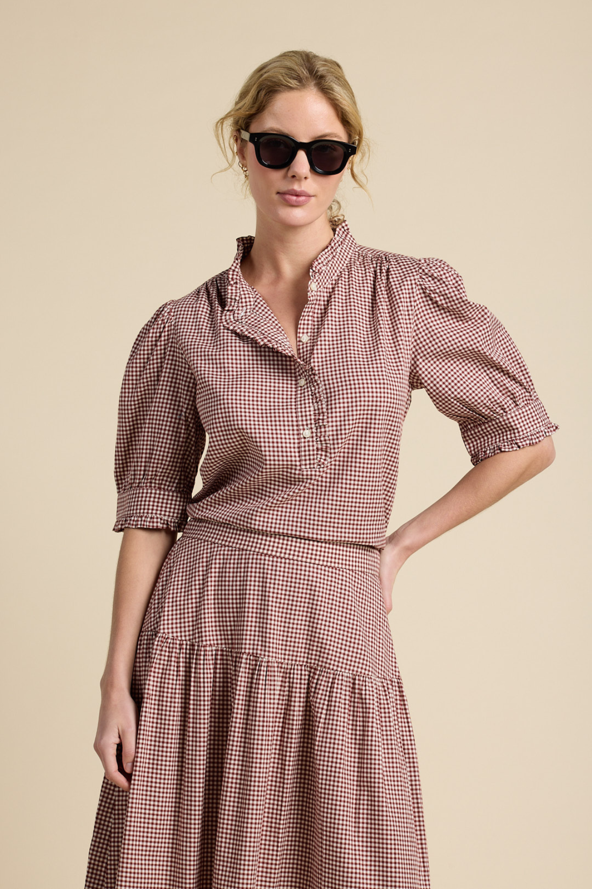 A woman modeling a brown and white gingham tiered dress with puff sleeves and a high collar, styled with black sunglasses against a solid beige background.