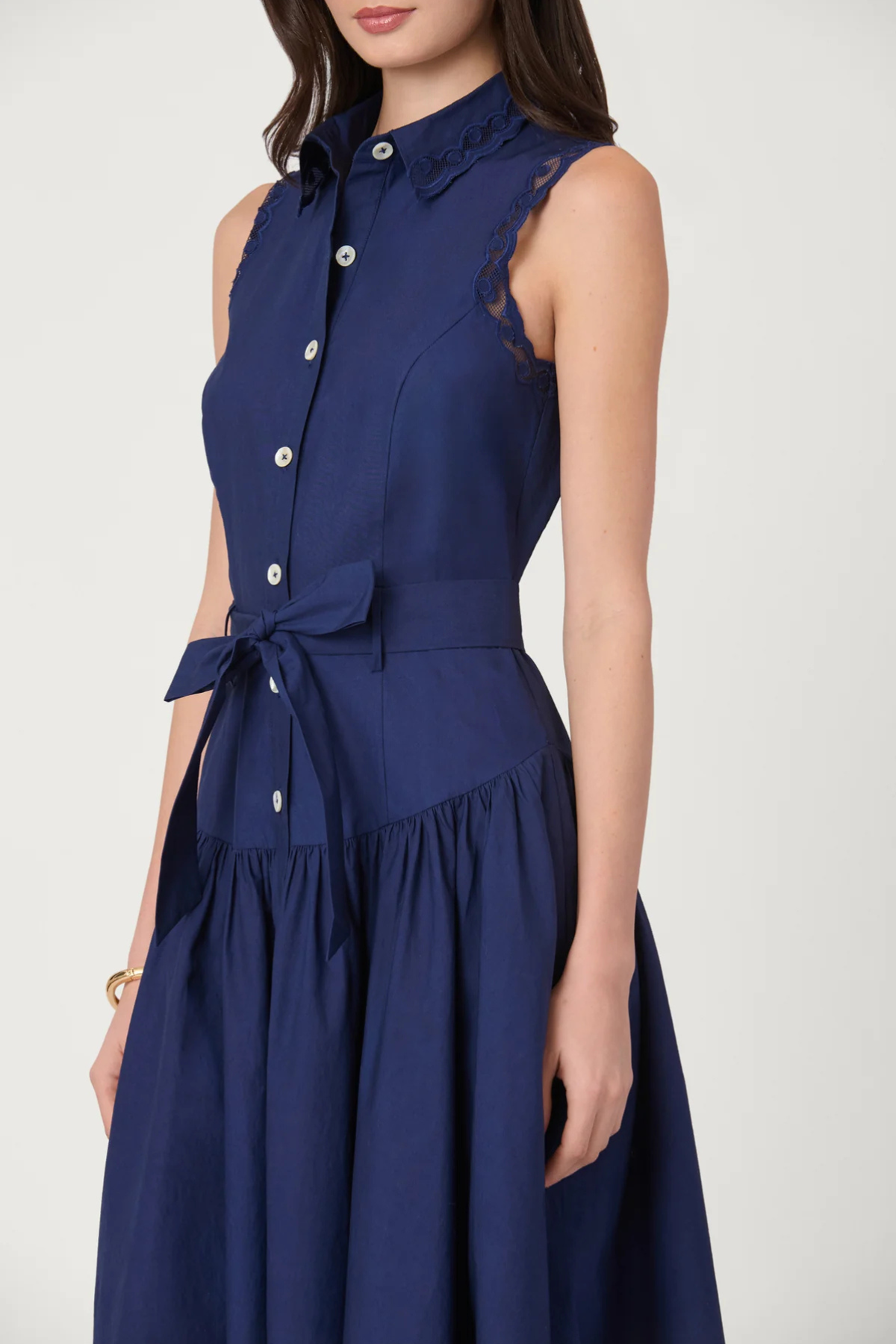 A sleeveless navy blue midi dress with a high collar, button-front bodice, matching tie belt, and lace trim details, shown on a model against a light gray background.