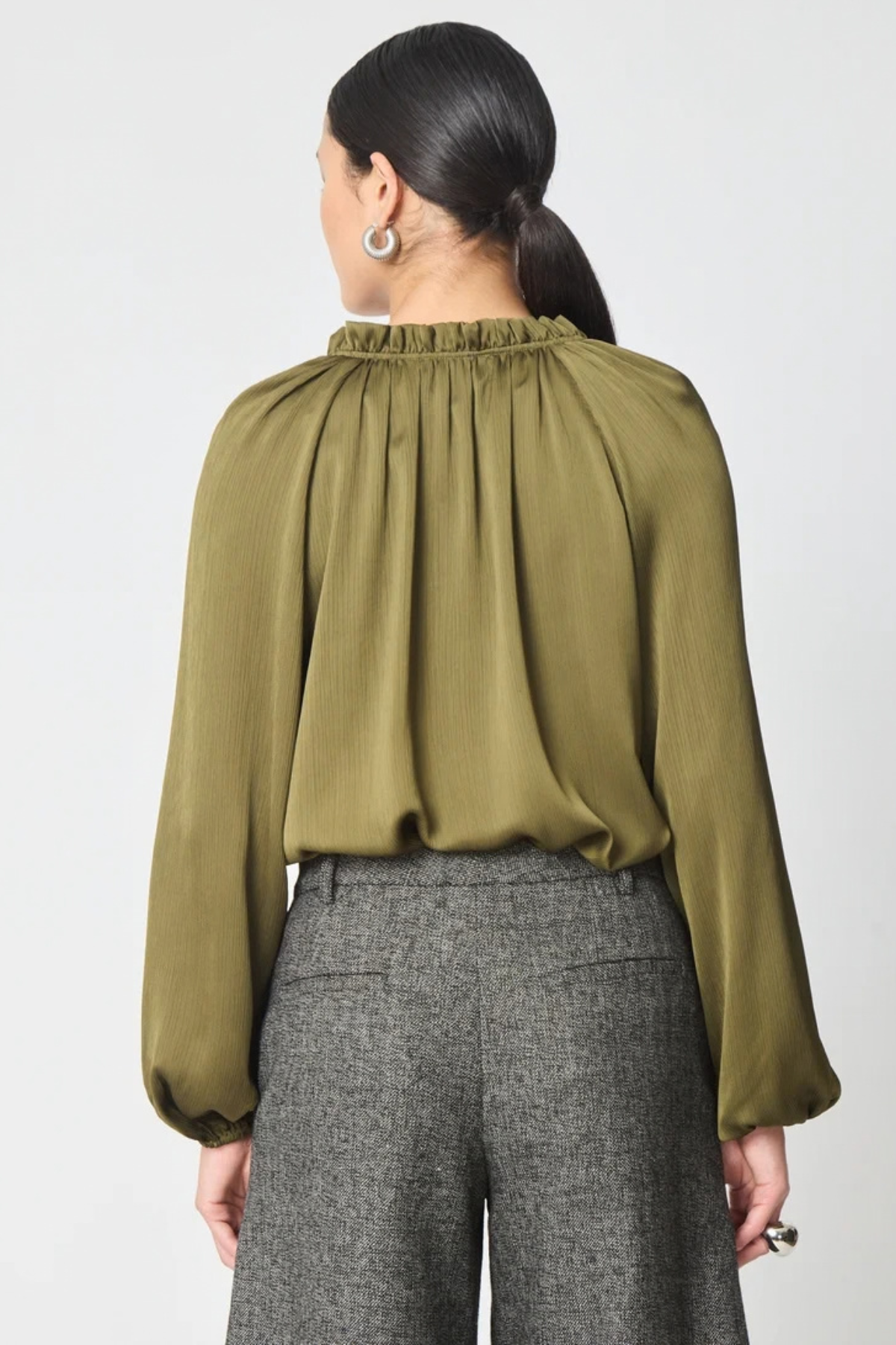 A woman shown from the back wearing a long-sleeve olive green blouse with a gathered, pleated high neckline and grey textured trousers, against a plain light gray background.