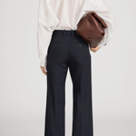 A model shown from the back wearing high-waisted navy wide-leg trousers with rolled cuffs, a white sheer long-sleeve blouse, and holding a brown leather clutch bag against a light gray studio background.