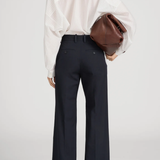 A model shown from the back wearing high-waisted navy wide-leg trousers with rolled cuffs, a white sheer long-sleeve blouse, and holding a brown leather clutch bag against a light gray studio background.