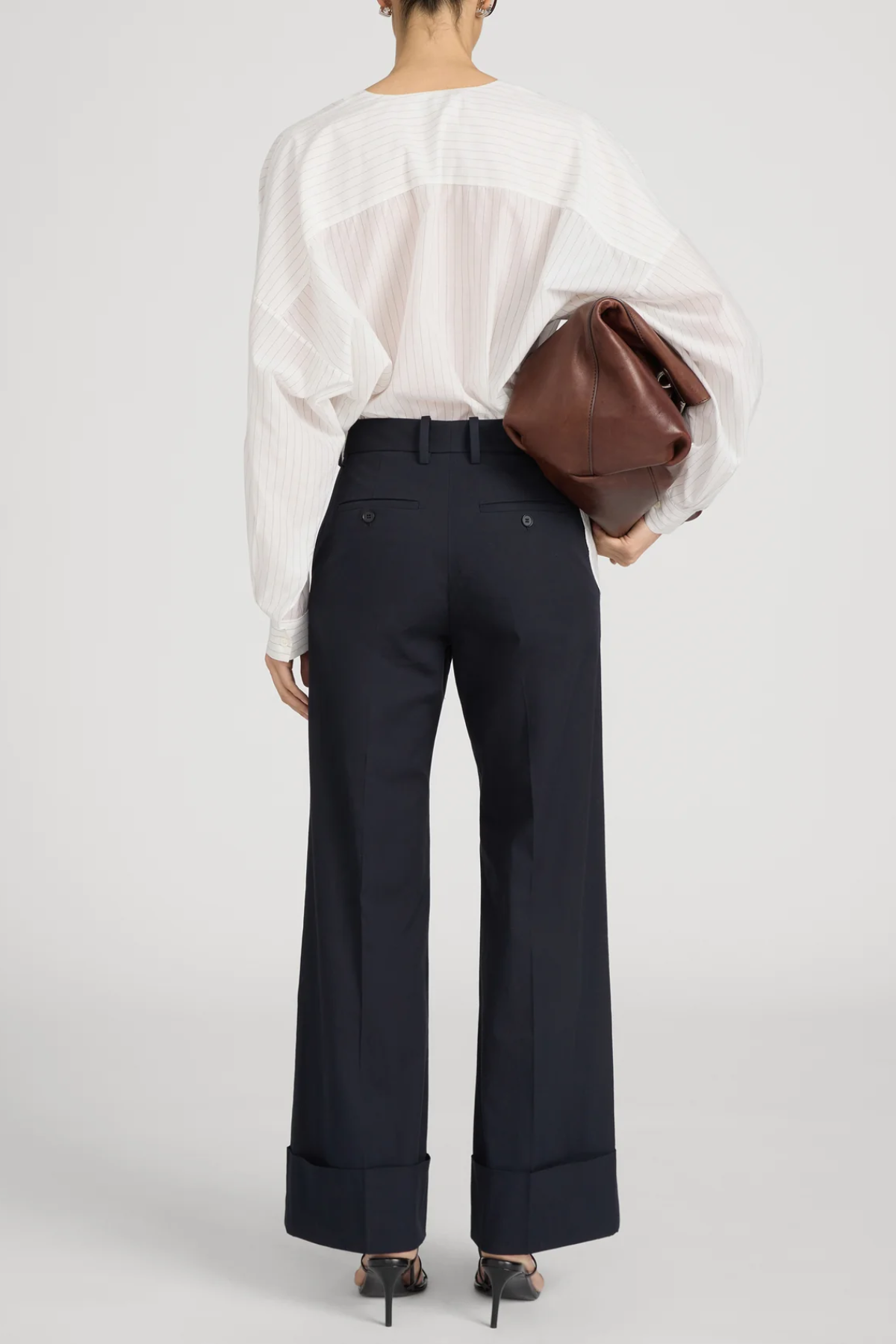 A model shown from the back wearing high-waisted navy wide-leg trousers with rolled cuffs, a white sheer long-sleeve blouse, and holding a brown leather clutch bag against a light gray studio background.