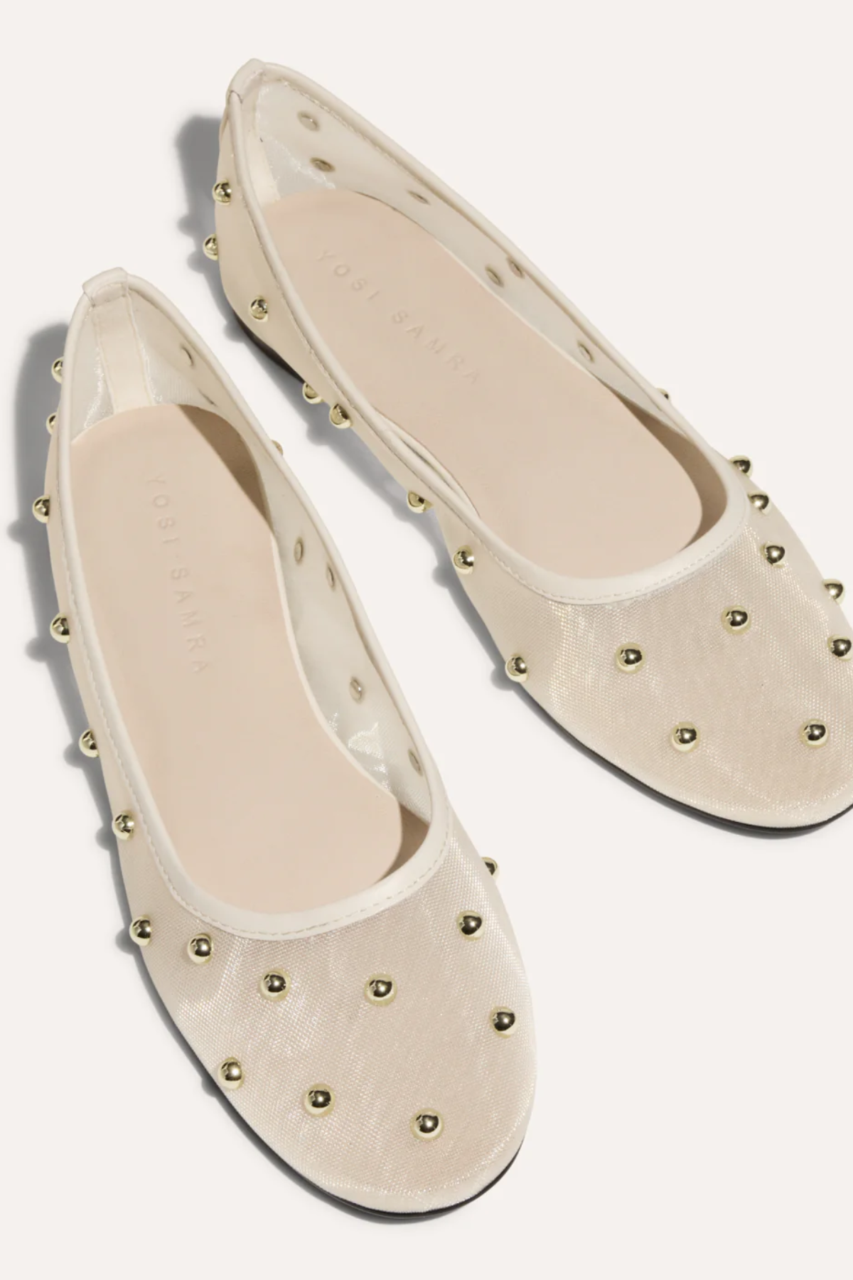 A pair of cream-colored ballet flats with a textured fabric upper and gold-tone metal studs, shown from a top-down angle on a light beige background.