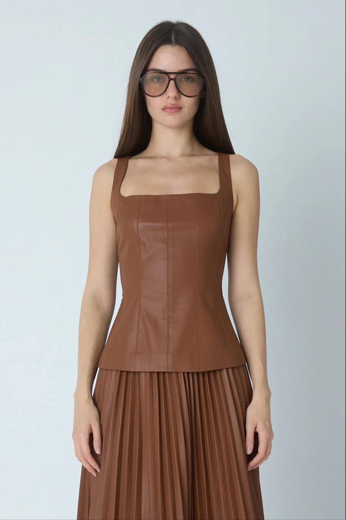 A woman modeling a brown faux-leather sleeveless top with a square neckline and matching pleated skirt against a light gray background.