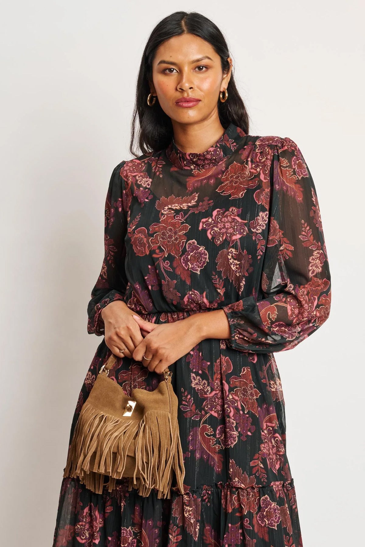 Woman wearing a floral dress holding a brown fringed bag against a white background
