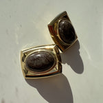 A pair of gold-toned rectangular cufflinks with oval dark brown mottled stone centers, displayed on a light off-white surface with strong sunlight and shadows.