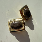 A pair of gold-toned rectangular cufflinks with oval dark brown mottled stone centers, displayed on a light off-white surface with strong sunlight and shadows.