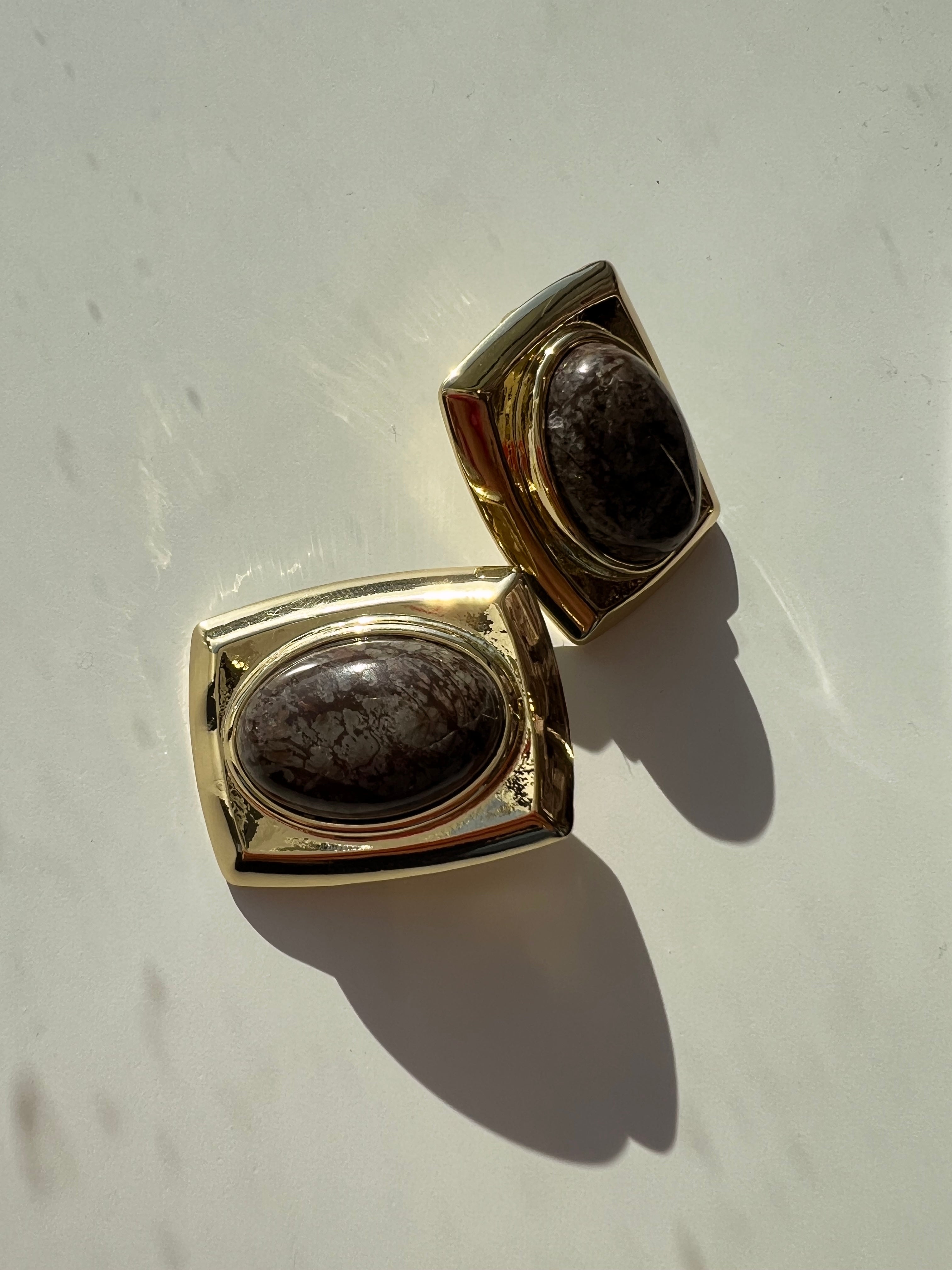 A pair of gold-toned rectangular cufflinks with oval dark brown mottled stone centers, displayed on a light off-white surface with strong sunlight and shadows.