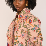 A woman with a large black afro is shown from the chest up, wearing a light peach-pink short-sleeve blouse with a colorful floral and botanical print. She is wearing gold hoop earrings against a plain, light off-white background.