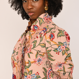 A woman with a large black afro is shown from the chest up, wearing a light peach-pink short-sleeve blouse with a colorful floral and botanical print. She is wearing gold hoop earrings against a plain, light off-white background.