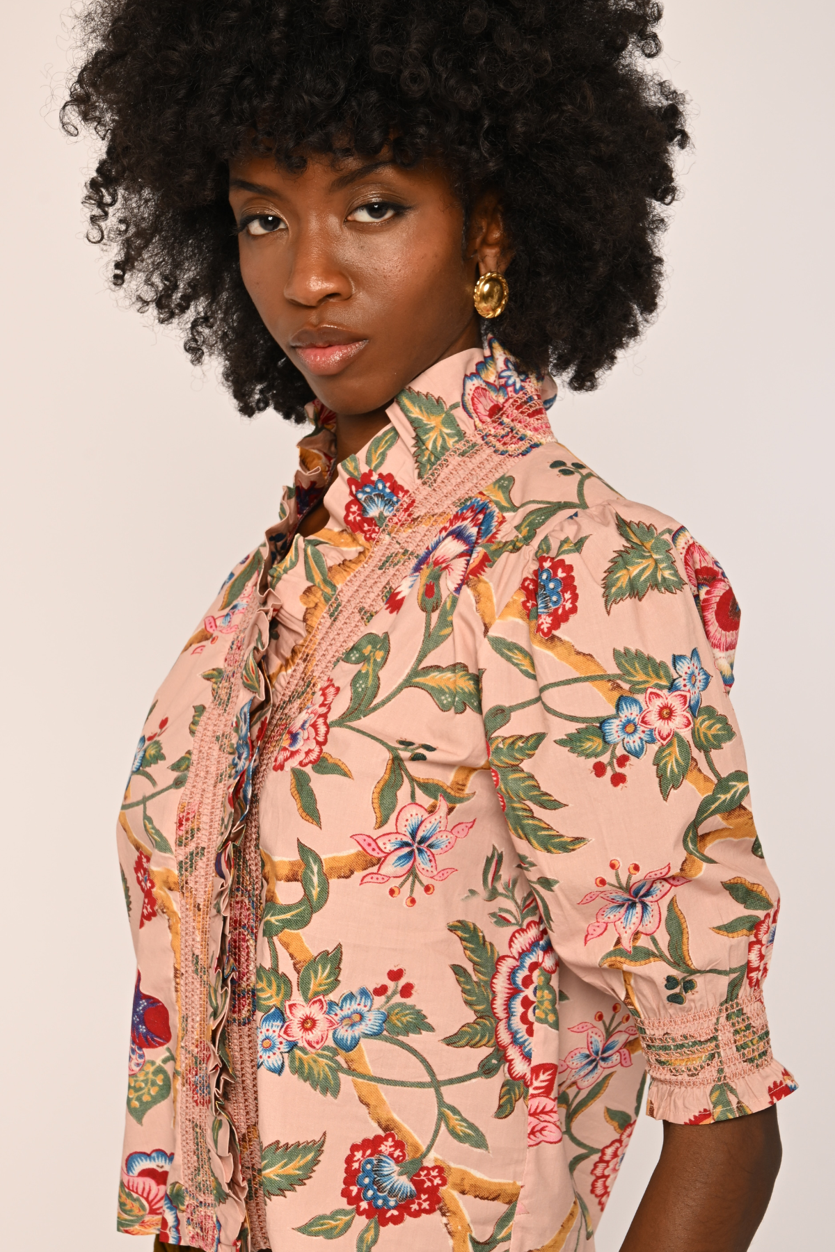 A woman with a large black afro is shown from the chest up, wearing a light peach-pink short-sleeve blouse with a colorful floral and botanical print. She is wearing gold hoop earrings against a plain, light off-white background.