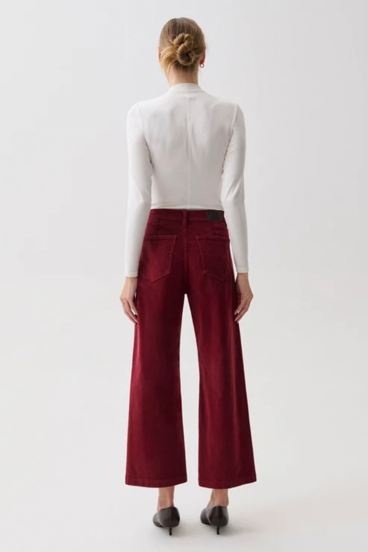 A model shown from the back wearing a white long-sleeve mock neck top and high-waisted, wide-leg burgundy corduroy trousers, paired with black pointed-toe heels, against a plain white studio background.