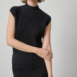 A woman wearing a black ribbed knit mock-neck top with cap sleeves and a matching ribbed knit skirt, photographed against a plain light gray studio background.