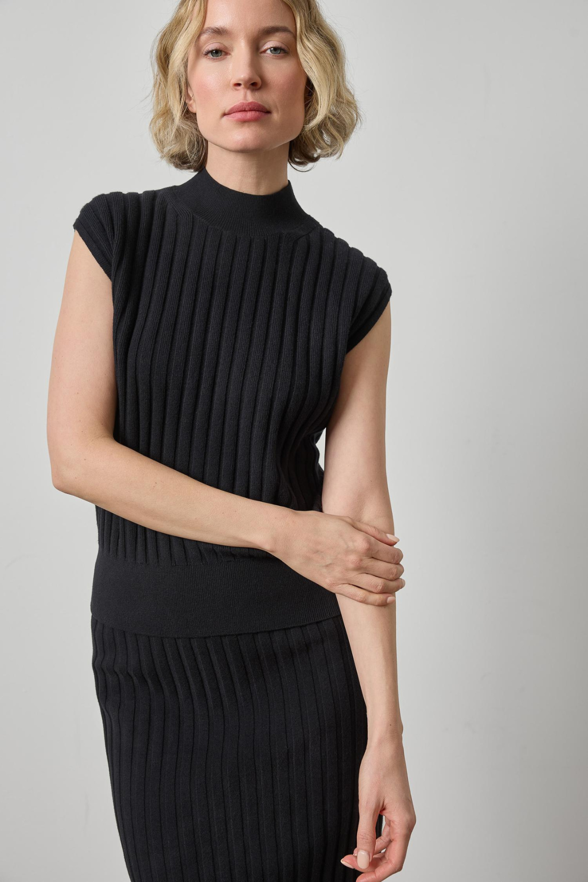 A woman wearing a black ribbed knit mock-neck top with cap sleeves and a matching ribbed knit skirt, photographed against a plain light gray studio background.