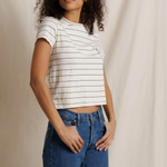 A woman with curly dark hair wearing a white short-sleeve crop top with thin black horizontal stripes and blue jeans, standing against a draped off-white fabric backdrop.