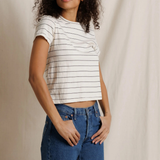 A woman with curly dark hair wearing a white short-sleeve crop top with thin black horizontal stripes and blue jeans, standing against a draped off-white fabric backdrop.