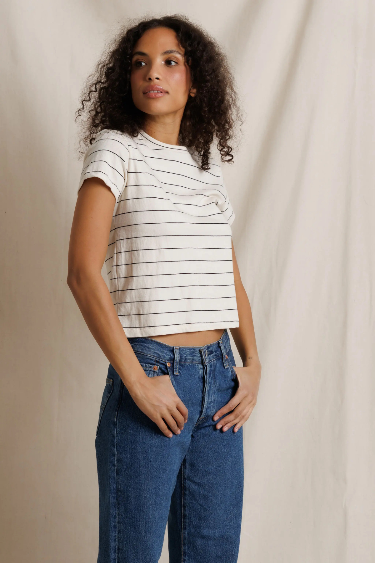 A woman with curly dark hair wearing a white short-sleeve crop top with thin black horizontal stripes and blue jeans, standing against a draped off-white fabric backdrop.