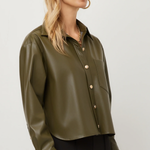 A woman wearing a cropped, long-sleeve olive green faux-leather button-up shirt with gold-tone buttons, paired with black pants, against a plain light gray studio background.