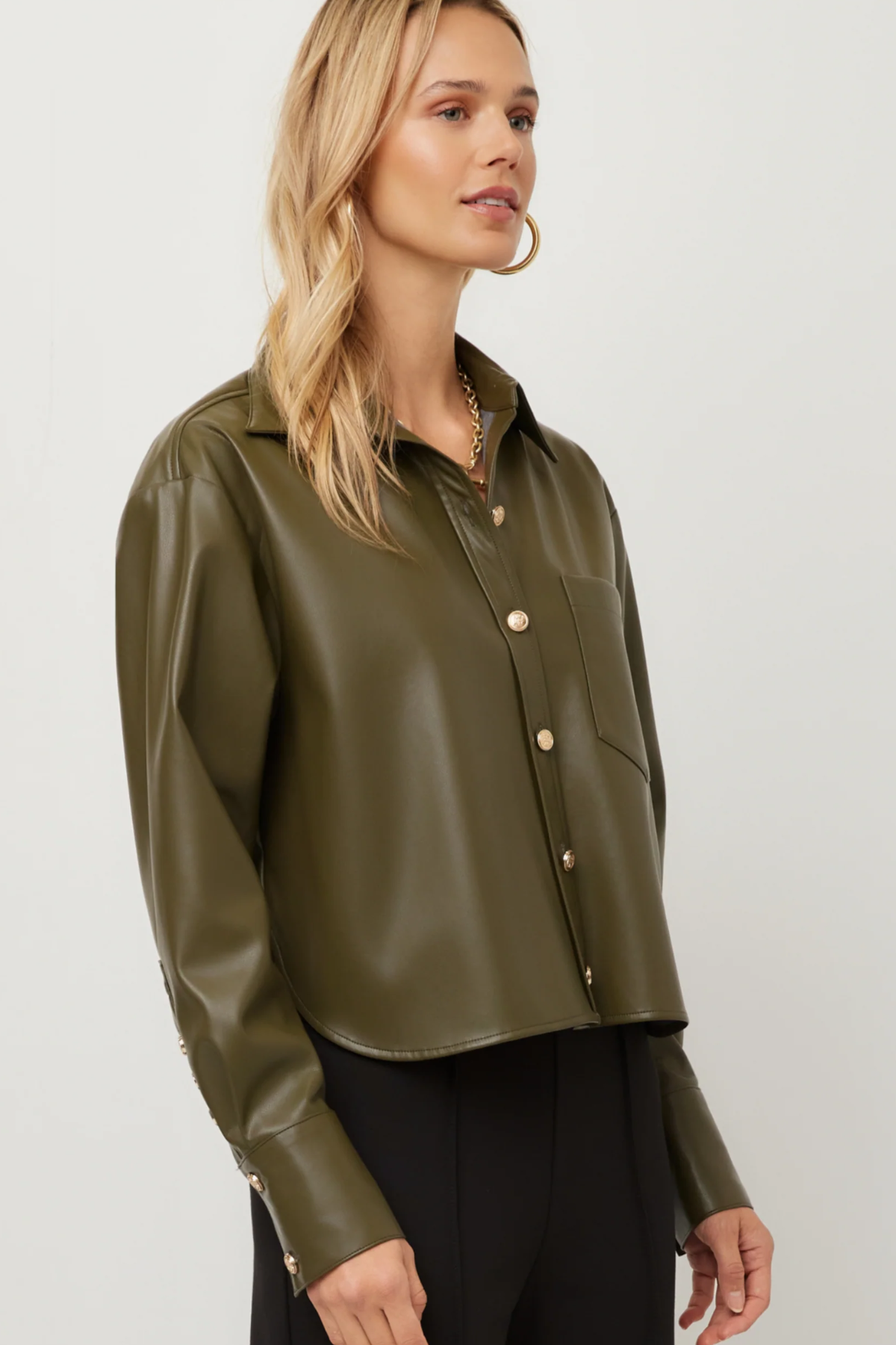 A woman wearing a cropped, long-sleeve olive green faux-leather button-up shirt with gold-tone buttons, paired with black pants, against a plain light gray studio background.