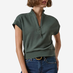A woman modeling a dark green short-sleeve quarter-zip knit top with a ribbed hem, paired with blue jeans, against a plain light gray background.