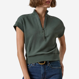 A woman modeling a dark green short-sleeve quarter-zip knit top with a ribbed hem, paired with blue jeans, against a plain light gray background.