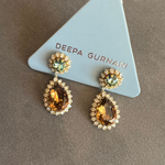 A pair of gold drop earrings with teardrop-shaped amber gemstones surrounded by small clear stones, and round green gemstones at the top, displayed on a light blue triangular card printed with the brand name 'DEEPA GURNANI'.
