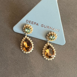 A pair of gold drop earrings with teardrop-shaped amber gemstones surrounded by small clear stones, and round green gemstones at the top, displayed on a light blue triangular card printed with the brand name 'DEEPA GURNANI'.