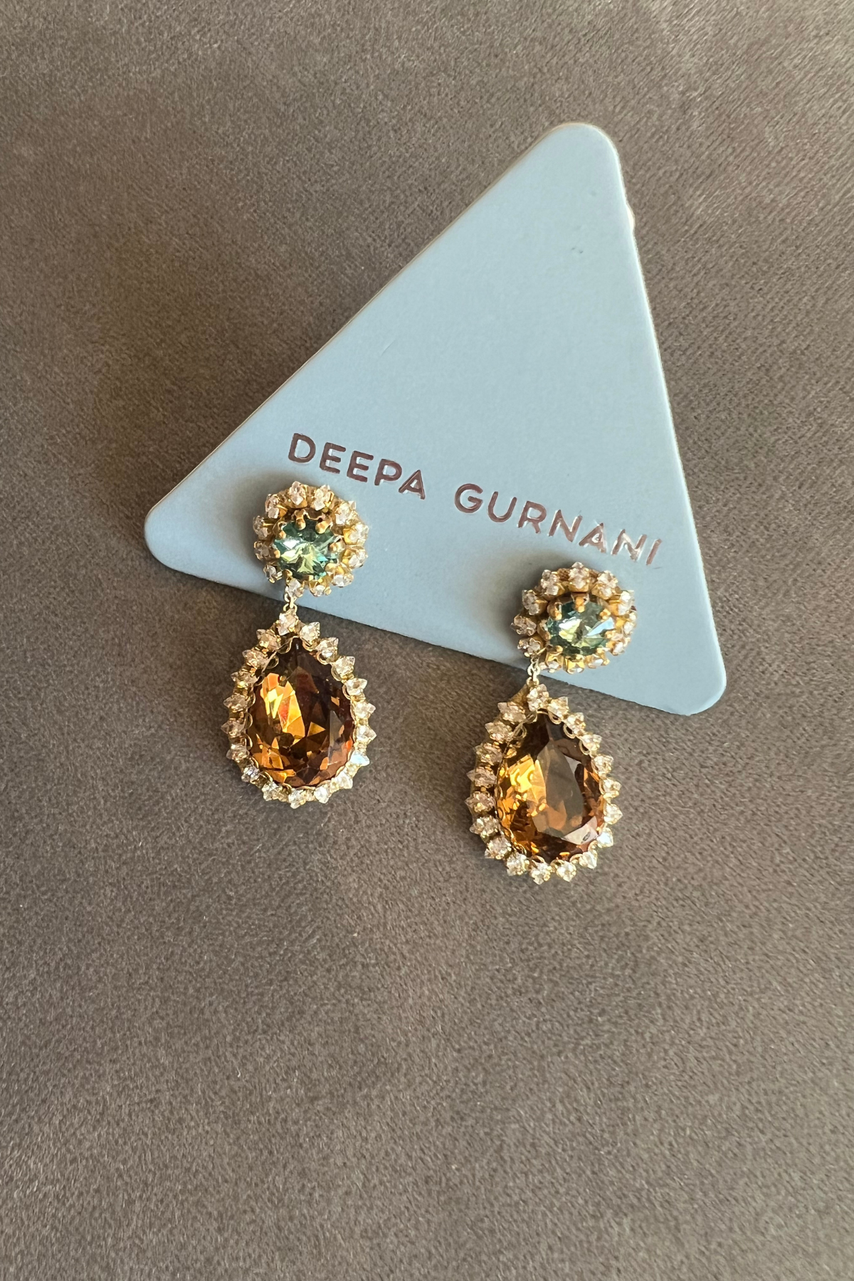 A pair of gold drop earrings with teardrop-shaped amber gemstones surrounded by small clear stones, and round green gemstones at the top, displayed on a light blue triangular card printed with the brand name 'DEEPA GURNANI'.