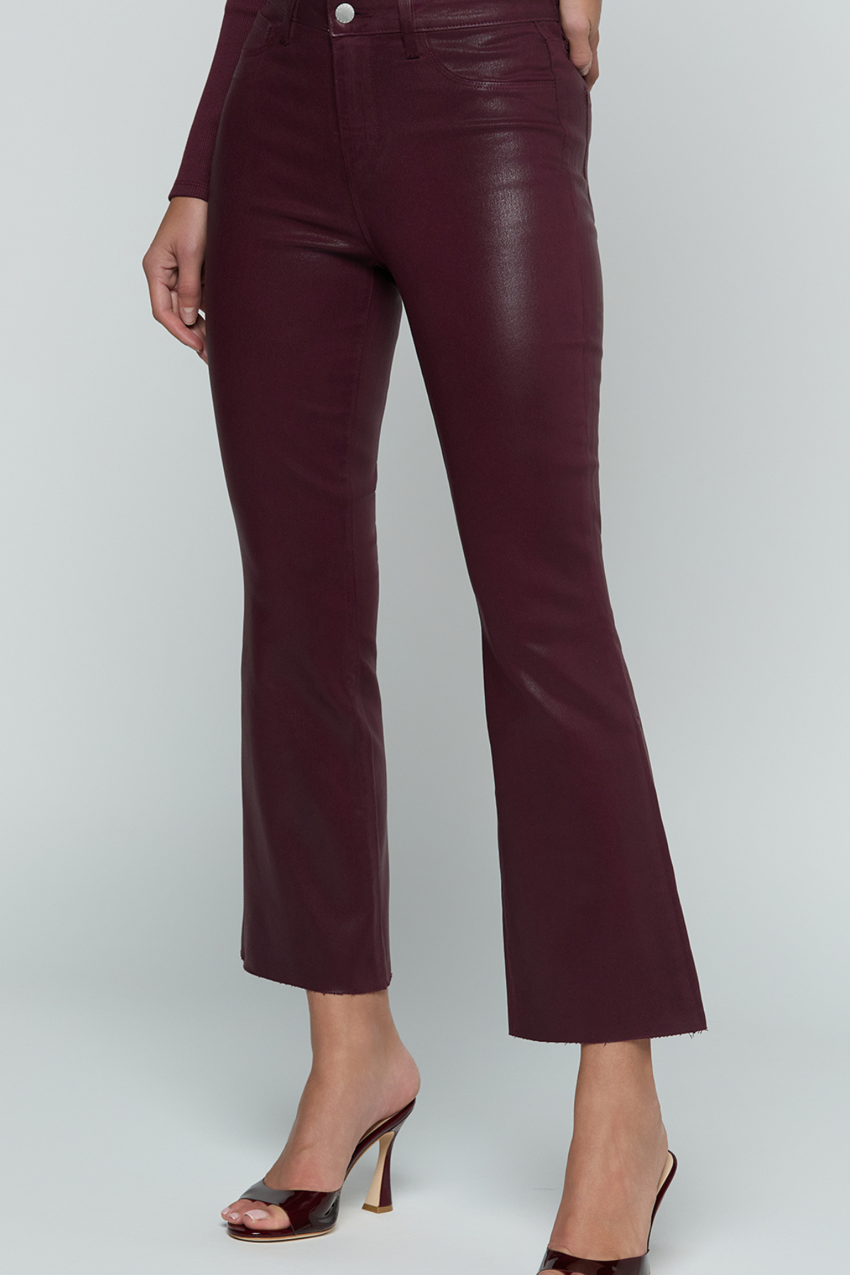 A model wearing dark burgundy faux-leather cropped flare pants with a silver button closure, paired with dark brown heeled sandals, against a light gray studio background.