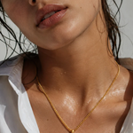 A close-up lifestyle shot of a woman wearing a gold coin pendant necklace, with water droplets on her skin and a white shirt collar visible.