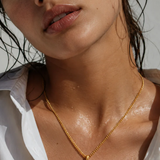 A close-up lifestyle shot of a woman wearing a gold coin pendant necklace, with water droplets on her skin and a white shirt collar visible.