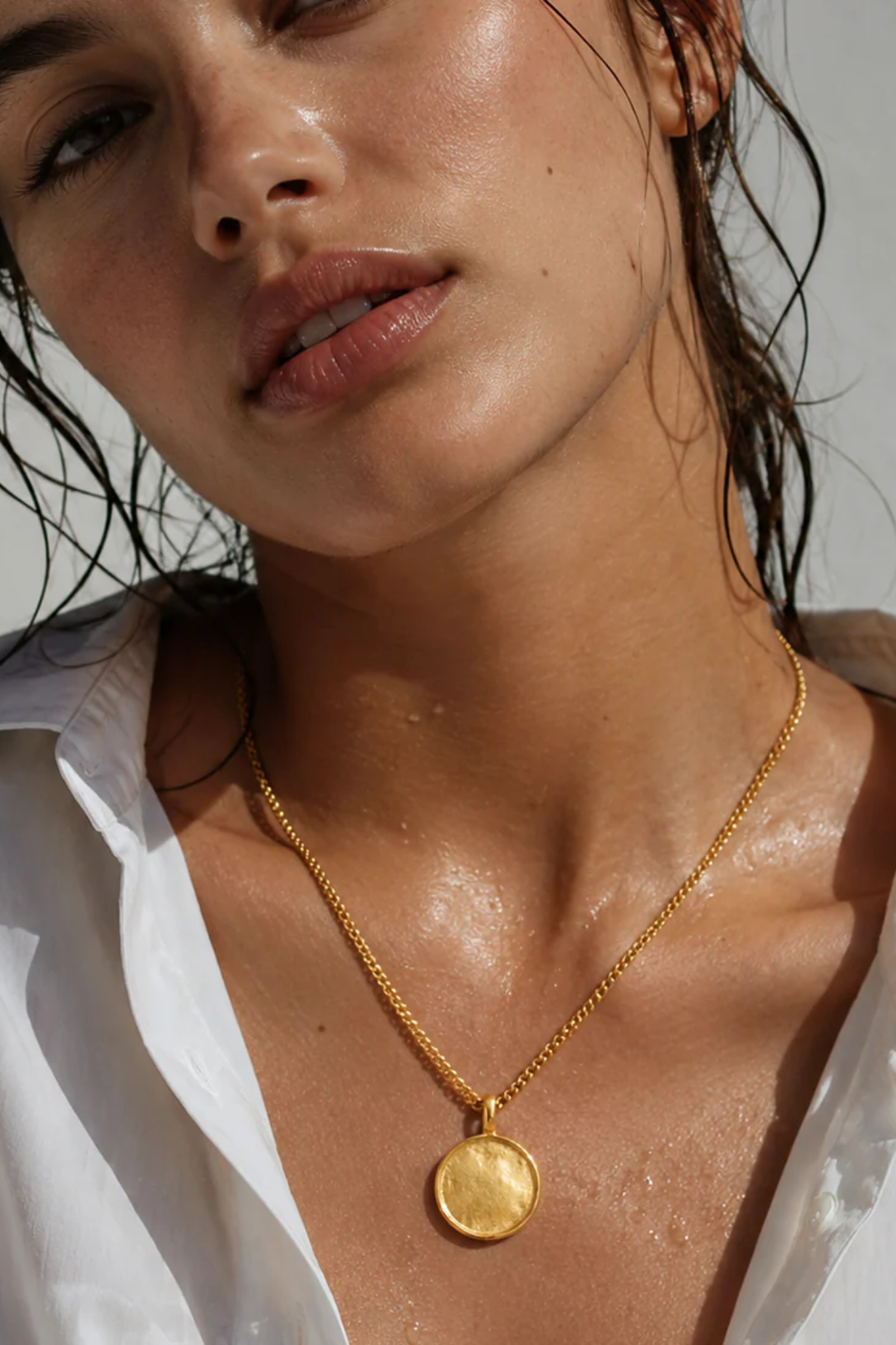 A close-up lifestyle shot of a woman wearing a gold coin pendant necklace, with water droplets on her skin and a white shirt collar visible.