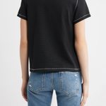 A woman shown from the back wearing a black short-sleeve t-shirt with white contrast stitching and blue jeans, against a plain white studio background.