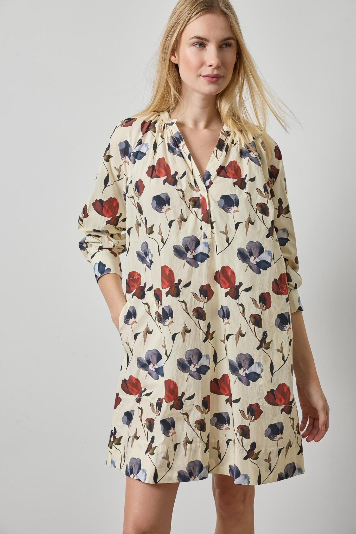 A woman modeling a cream-colored long-sleeve shirt dress with a red and navy blue floral print, set against a plain light gray studio background.