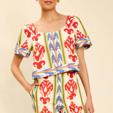 A woman modeling a matching two-piece printed outfit with short sleeves and wide-leg pants. The set features a cream base with vertical stripes in green, blue, and red, interspersed with red floral motifs.