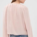 A woman shown from the back wearing a light pink long-sleeve blouse with a gathered, pleated neckline and flared sleeves, paired with blue jeans against a plain light gray studio background.