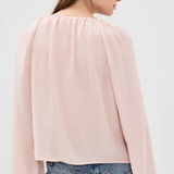 A woman shown from the back wearing a light pink long-sleeve blouse with a gathered, pleated neckline and flared sleeves, paired with blue jeans against a plain light gray studio background.