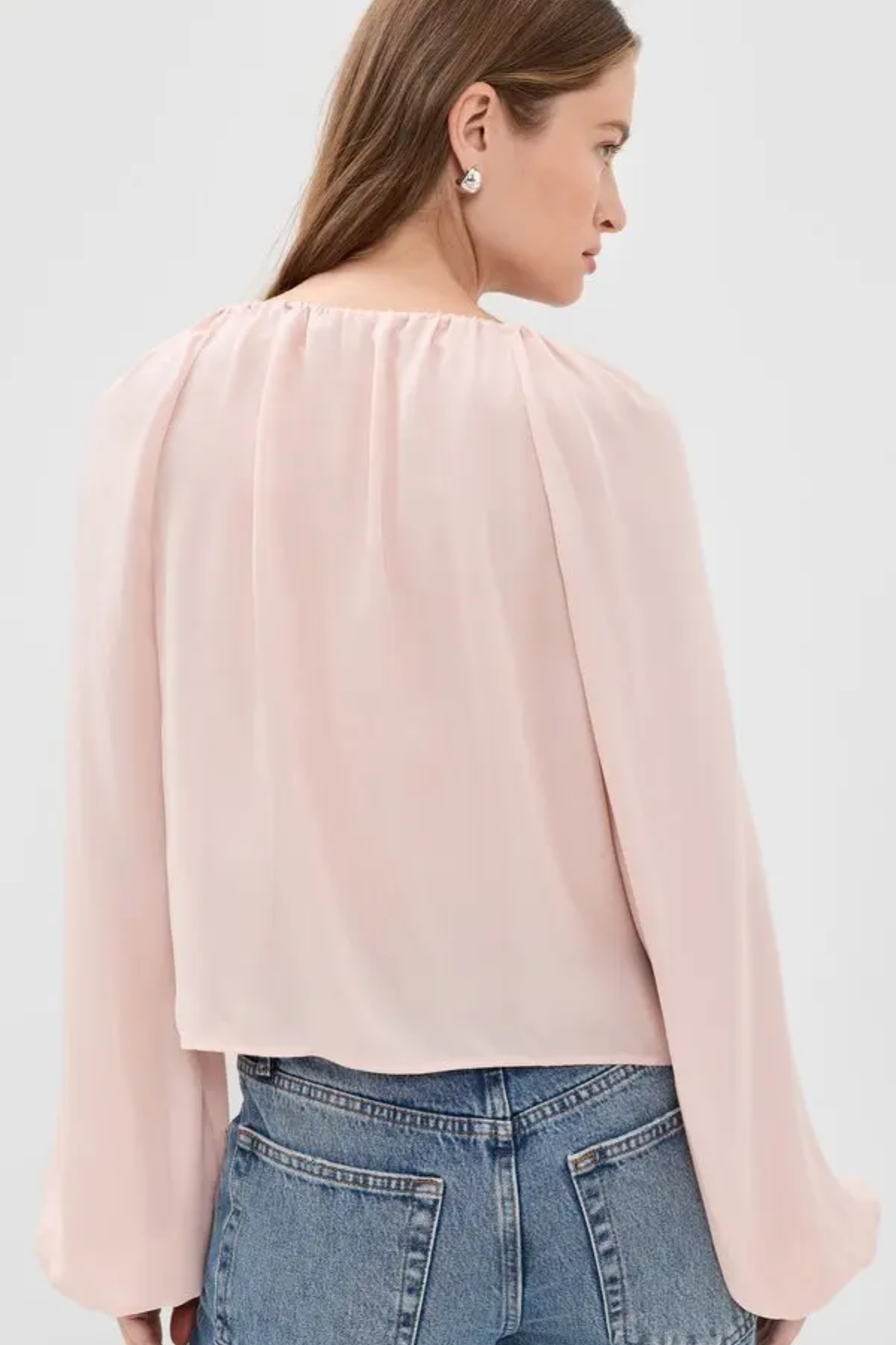 A woman shown from the back wearing a light pink long-sleeve blouse with a gathered, pleated neckline and flared sleeves, paired with blue jeans against a plain light gray studio background.