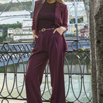 A woman models a matching burgundy three-piece suit consisting of a blazer, long-sleeve top, and wide-leg trousers, styled with a thin belt and burgundy sneakers, standing on a bridge with a river and buildings in the background.