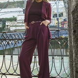 A woman models a matching burgundy three-piece suit consisting of a blazer, long-sleeve top, and wide-leg trousers, styled with a thin belt and burgundy sneakers, standing on a bridge with a river and buildings in the background.