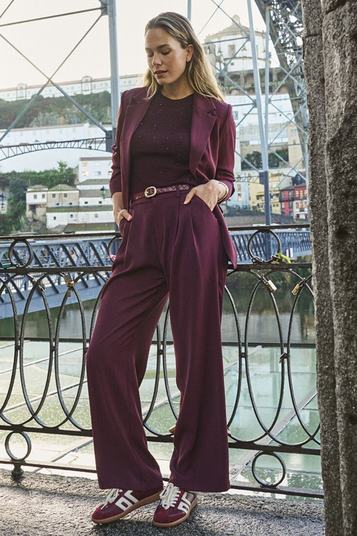A woman models a matching burgundy three-piece suit consisting of a blazer, long-sleeve top, and wide-leg trousers, styled with a thin belt and burgundy sneakers, standing on a bridge with a river and buildings in the background.