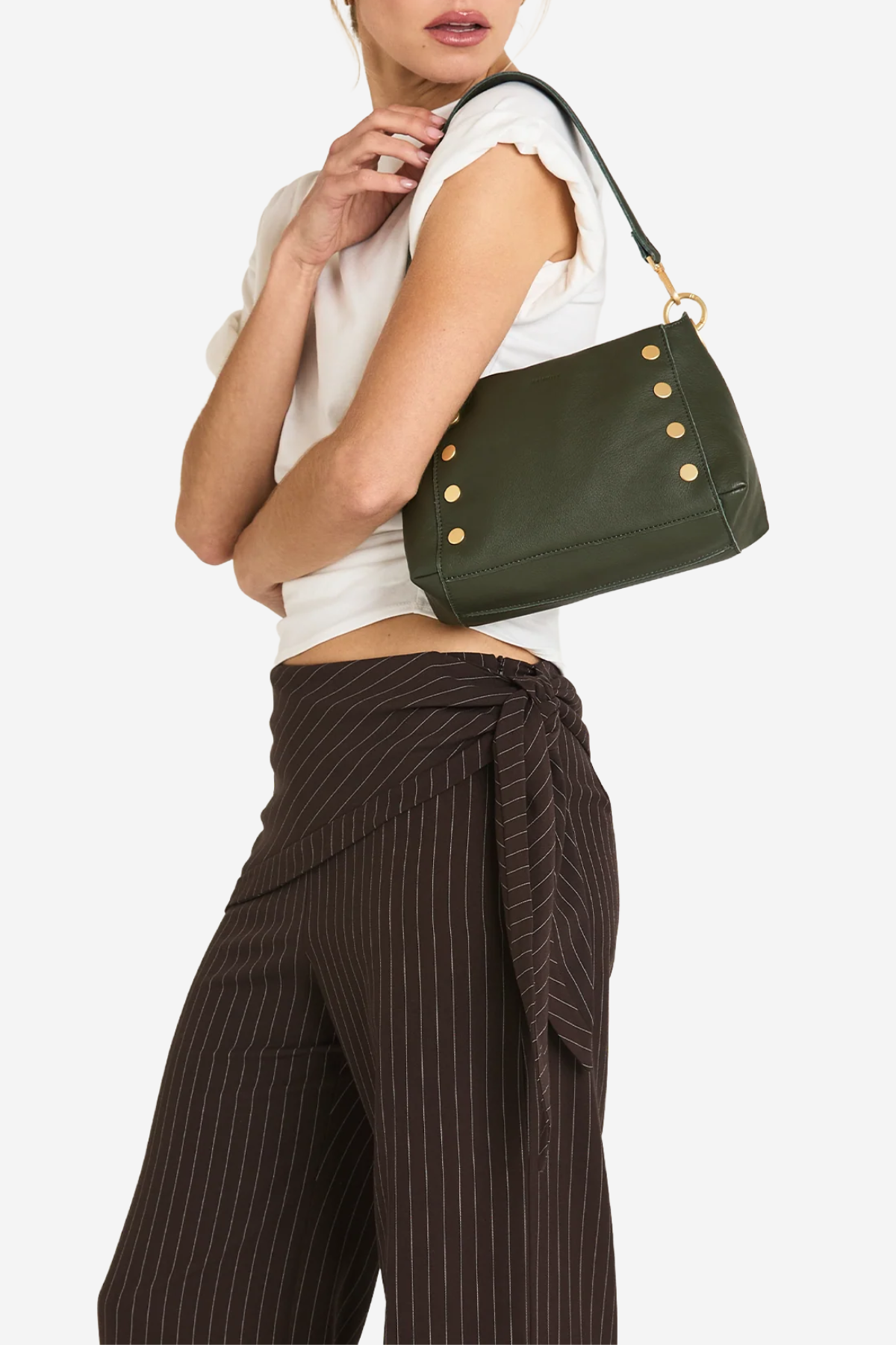 A model wearing a white sleeveless top and dark brown pinstriped wrap pants carries an olive green leather shoulder bag with gold-tone studs and a black strap against a light gray background.