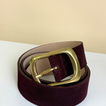 A dark burgundy velvet belt with a polished gold-tone metal buckle, coiled on a white surface against a beige background.