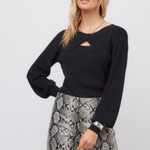 A woman modeling a black knit sweater with a chest cutout and long sleeves, paired with gray and black snakeskin print pants, against a light gray studio background.