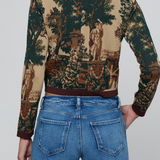 A cropped long-sleeve knit top with a classic toile landscape print in beige, green, and brown, featuring dark brown ribbed collar, cuffs, and hem, shown on a model from the back wearing blue jeans.