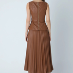 A woman viewed from the back wearing a sleeveless brown faux-leather top and matching pleated midi skirt, paired with black heeled boots, against a light gray studio background.