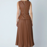 A woman viewed from the back wearing a sleeveless brown faux-leather top and matching pleated midi skirt, paired with black heeled boots, against a light gray studio background.