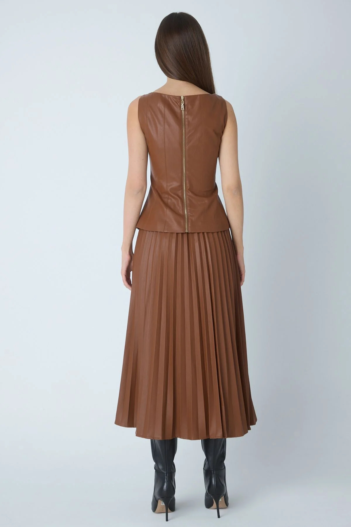 A woman viewed from the back wearing a sleeveless brown faux-leather top and matching pleated midi skirt, paired with black heeled boots, against a light gray studio background.