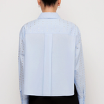 A model shown from the back wearing a light blue long-sleeve shirt with eyelet embroidery and black trousers, set against a plain white studio background.