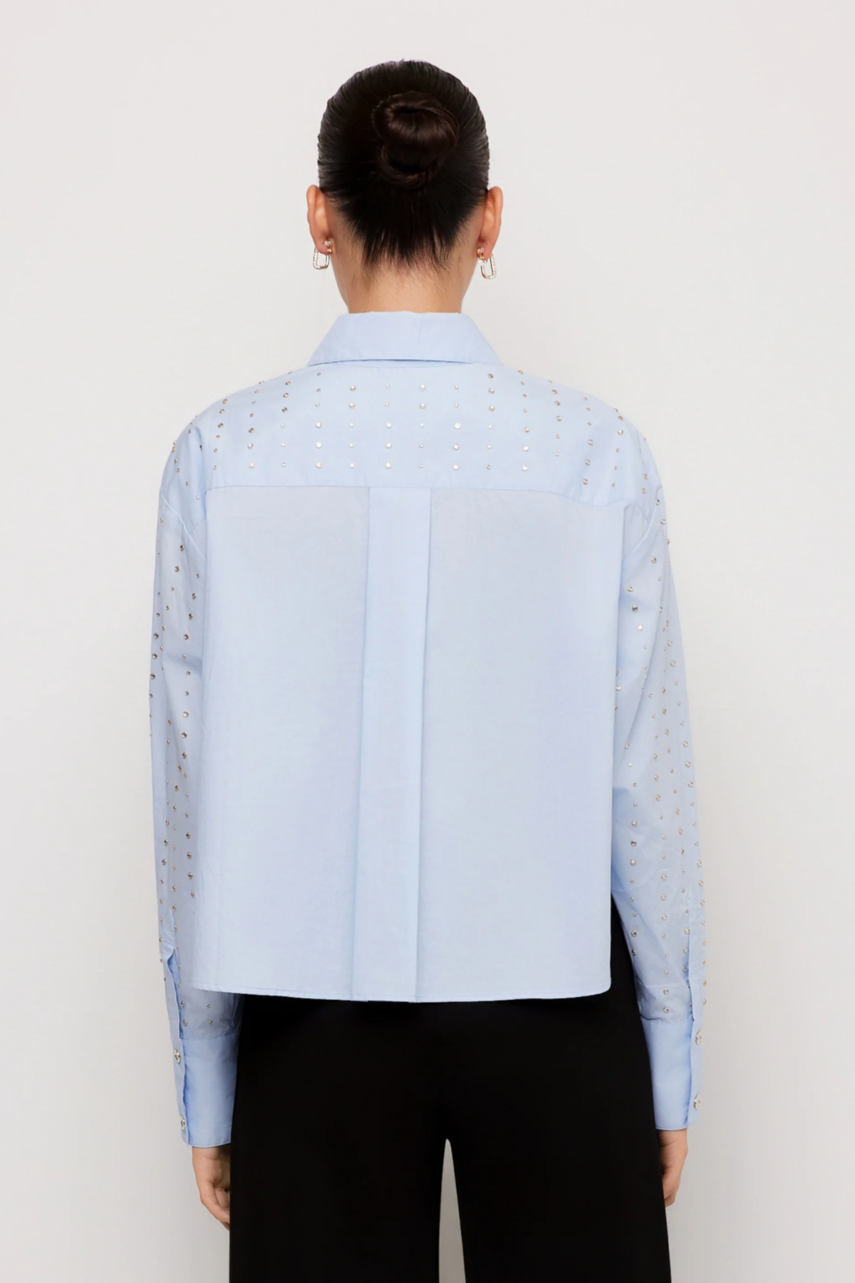A model shown from the back wearing a light blue long-sleeve shirt with eyelet embroidery and black trousers, set against a plain white studio background.